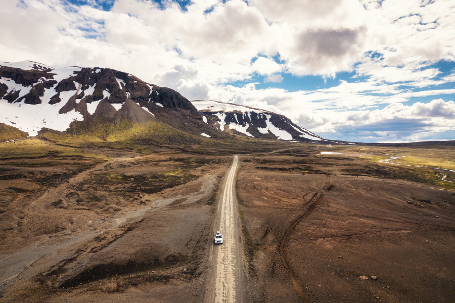 A campervan seen from above from a distance on a dirt road, surrounded by brown hills, then snow topped mountains and a cloudy sky in the background.