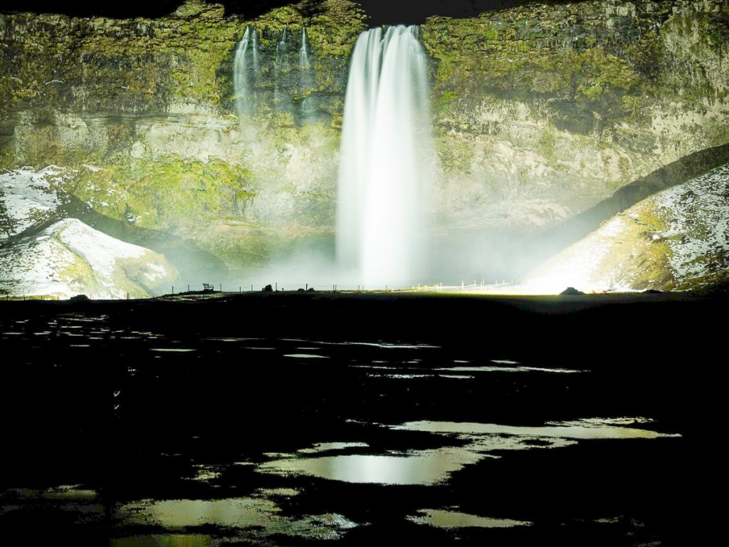 A stunning waterfall surrounded by cliffs lit up at night, with dark ground and puddles in the foreground.