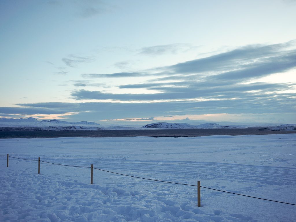 A snowy landscape leading to dark water and slightly cloudy sky.