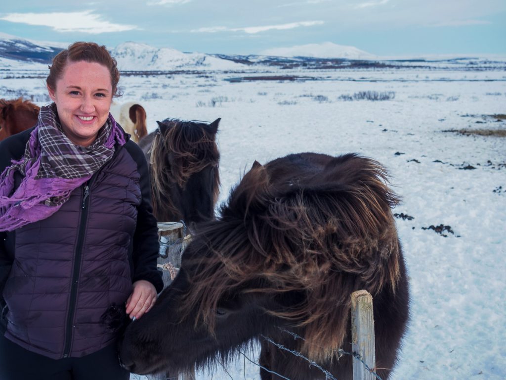 Sooz smiling at the camera while patting a pony in a snowy paddock with more ponies in the background.