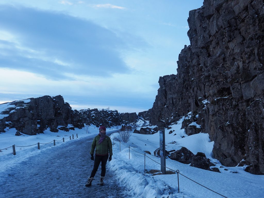 A woman rugged up in cold-weather gear standing on an icy road next to a black rocky cliff and snow.