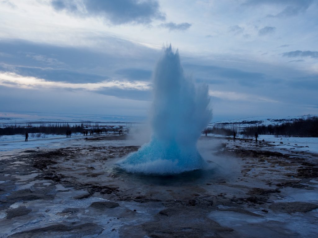 A geyser erupting out of rocky ground with snow in the background and a cloudy sky.