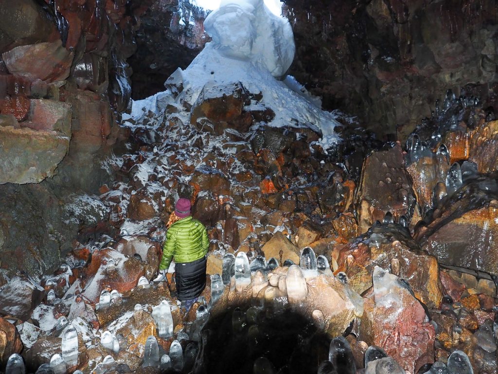 A woman rugged up in winter clothing standing inside a cave with unique rock and ice formations and snow near the top of the image.