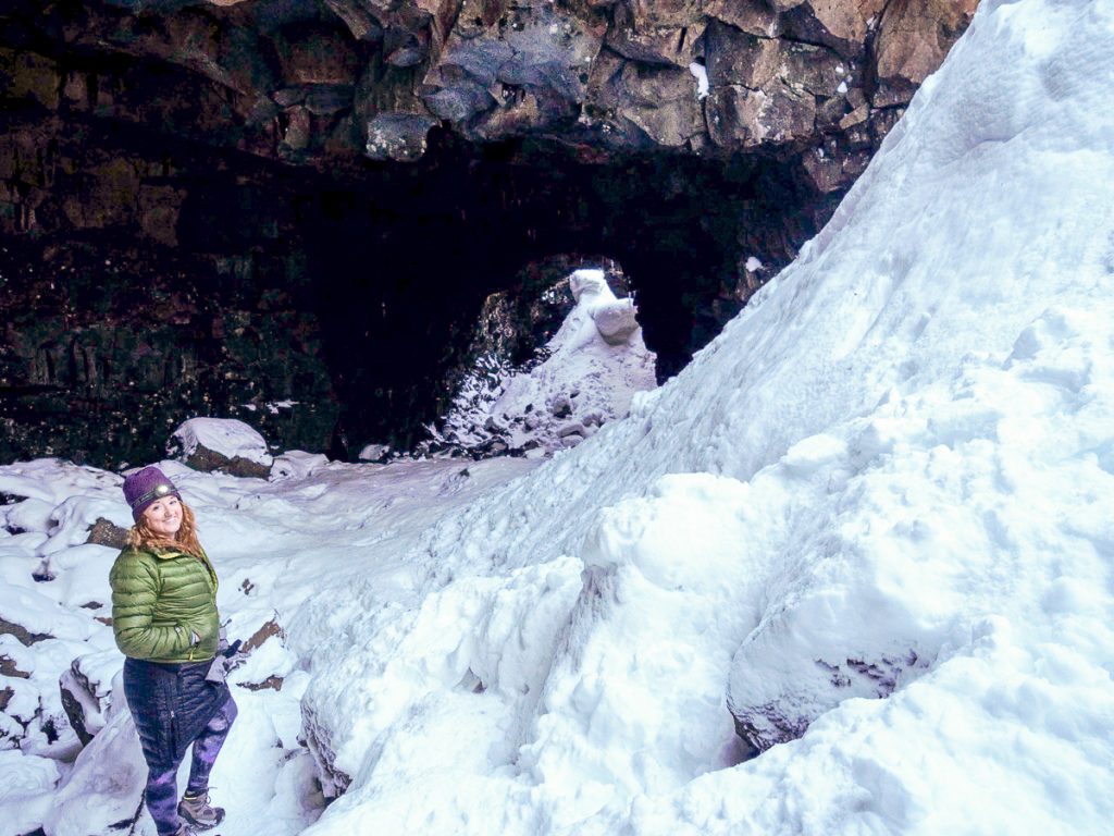 Sooz rugged up in winter gear standing at the opening of a rocky cave with snow all around.