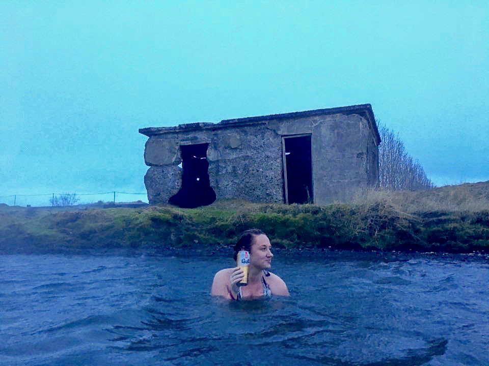 A woman in a lagoon holding a can with a stone building on the grass behind.