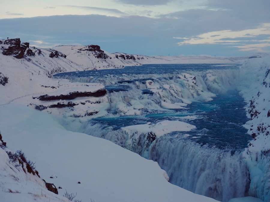 An icy river surrounded by snow and black rocks, with small waterfalls.