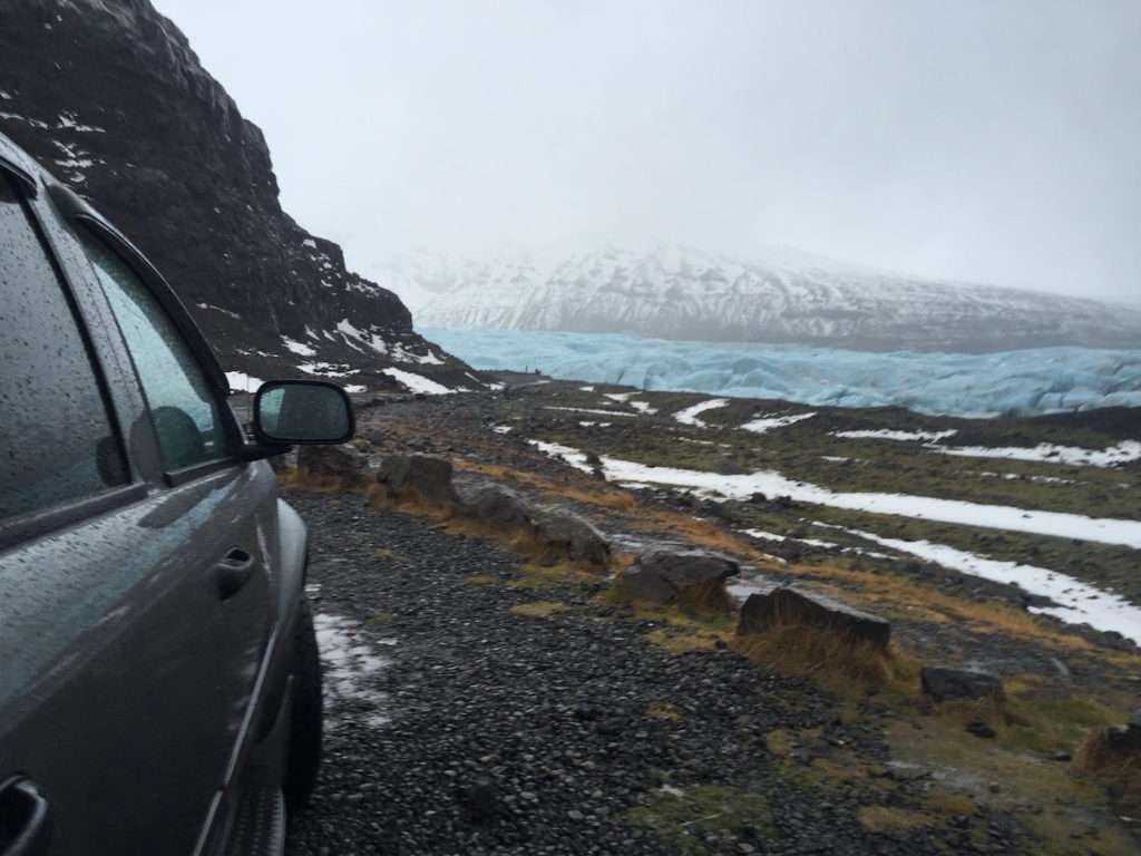 The front of a dark grey car seen from the side looking towards a landscape of snow, black rocks, ice and mist.