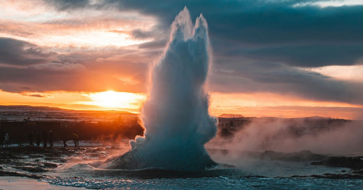 A geyser erupting with ice in the foreground and an orange sunset covered by clouds behind.