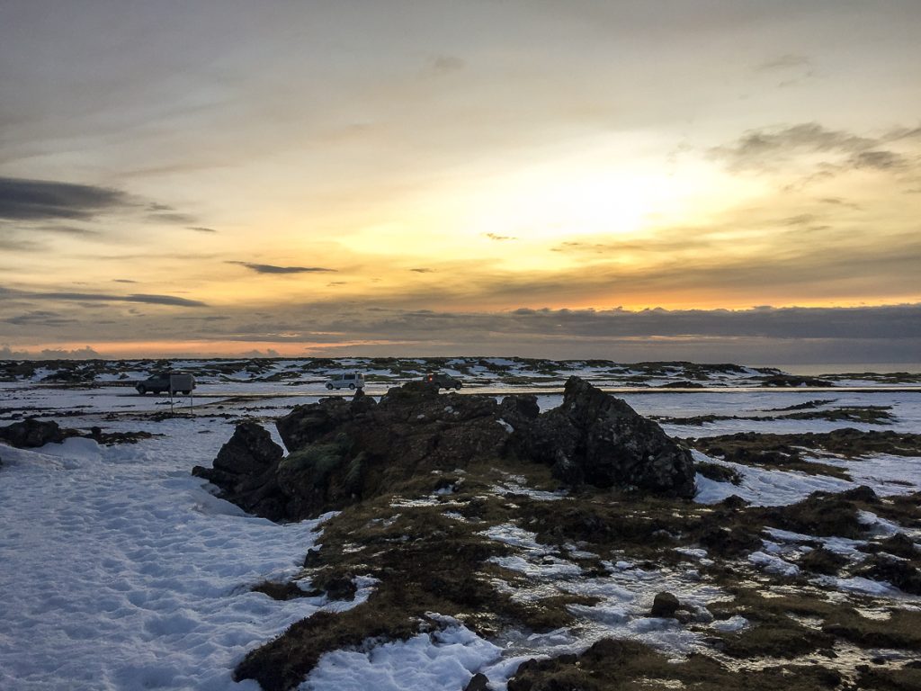 A rocky and snowy landscape with some cars and campervans in the background with a yellow sunset behind them.
