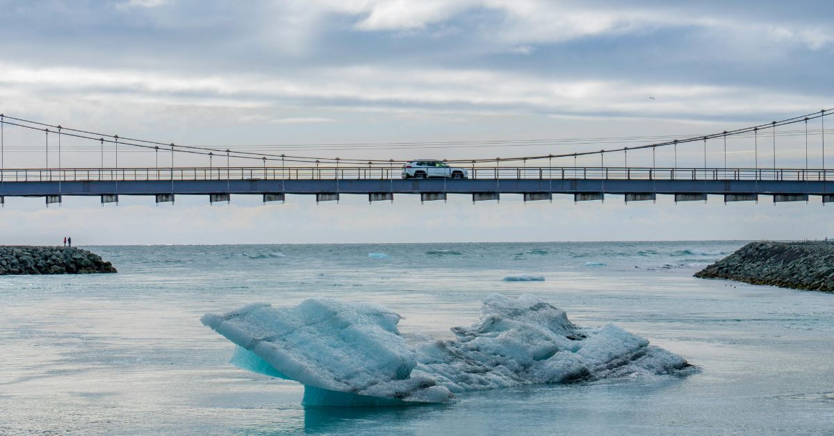 A car parked in the middle of a long bridge that's suspended over water with a large ice chunk in the foreground. In the background, two small figures stand on rock behind the bridge. The sky is cloudy.