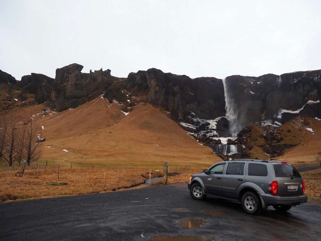 A dark grey campervan parked on gravel with a brown hill behind it leading to rocks and a small waterfall.