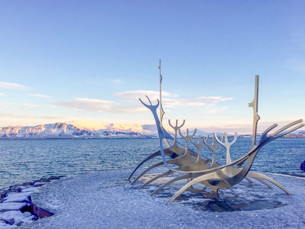 A metal sculpture that vaguely resembles a ship surrounded by icy ground with water and then snowy mountains and blue sky behind it.