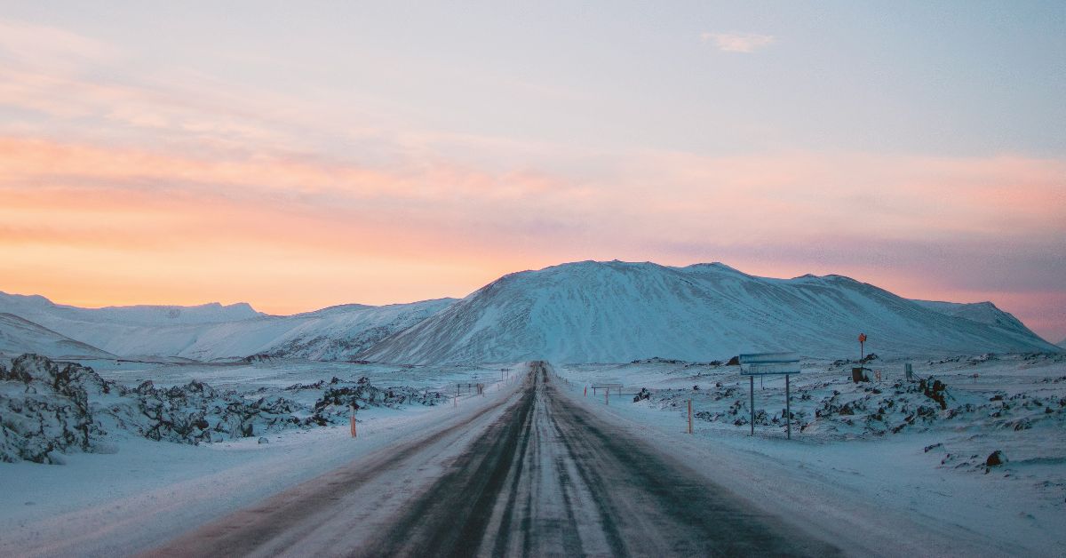 A snowy road in Iceland leading towards a hill and a pink sunset.