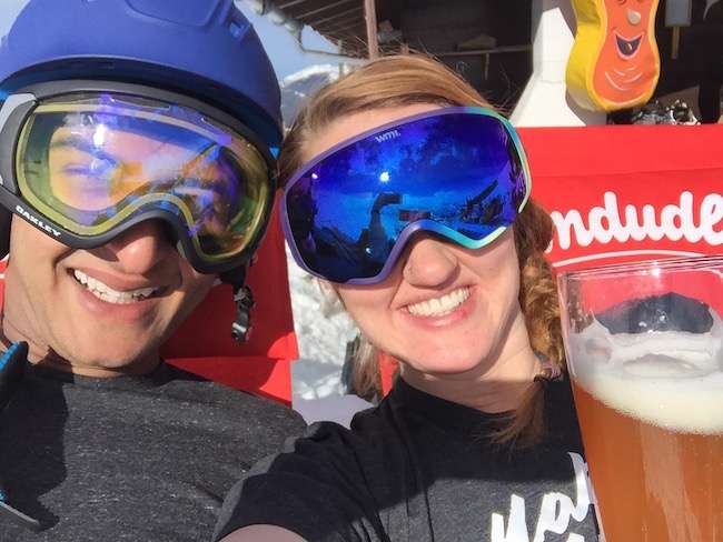 A man and woman wearing snow goggles grinning at the camera with a glass of beer in the foreground.