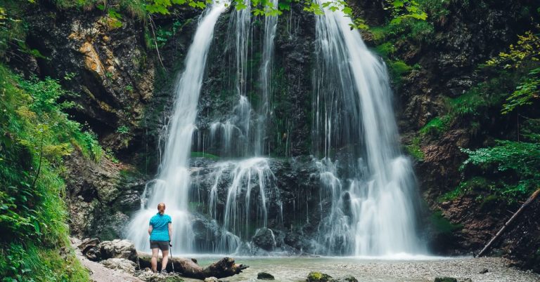 A woman leaning on a walking stick in front of a beautiful waterfall surrounded by greenery.