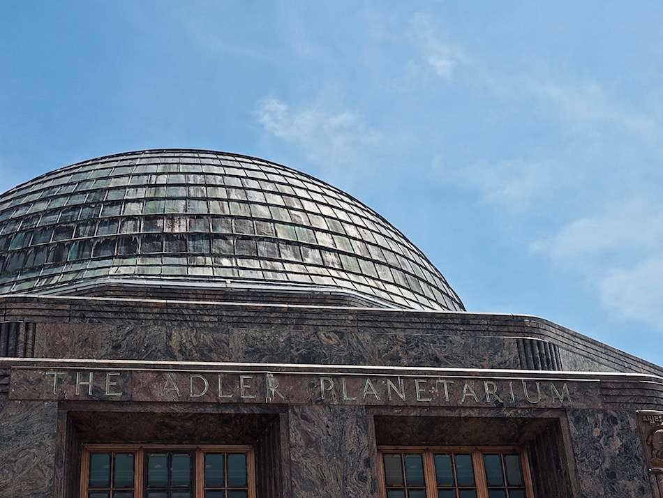 The domed roof above the entrance of a building with Adler Planetarium inscribed above the windows and a blue sky behind.