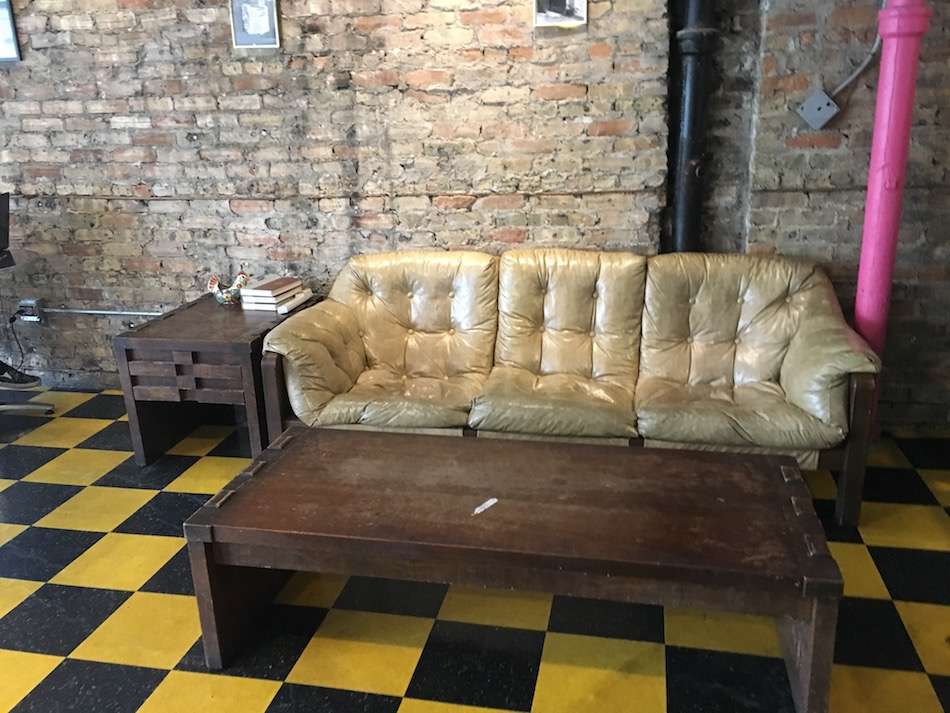 A light brown leather couch with a wooden coffee table in front of it on a checked yellow and black floor with brick walls behind.