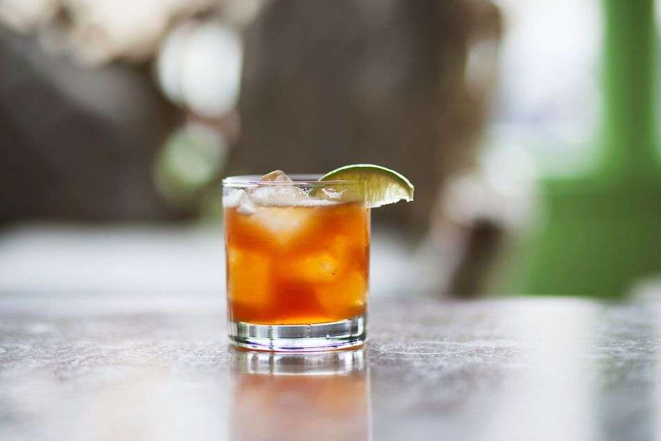 A close up of an orange drink with ice and a slice of lime in a short glass on a grey table.