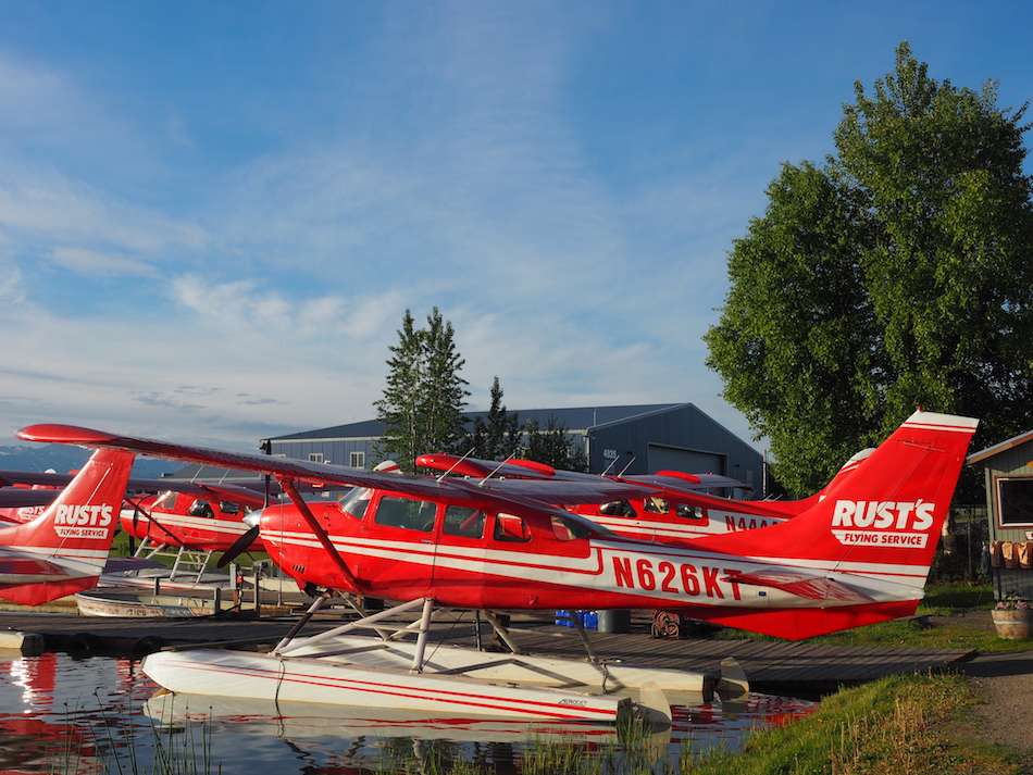 Some small red and white seaplanes on water in front of a building with a blue sky behind.