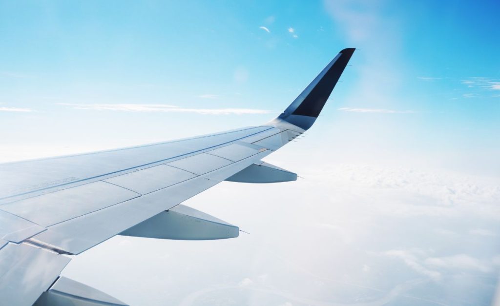 An airplane wing seen from a plane with fluffy clouds below and blue sky above.