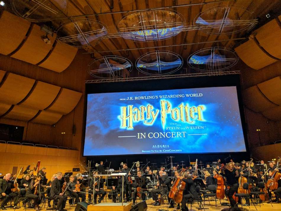 An orchestra sitting underneath a huge screen with text on it reading "Harry Potter in Concert."