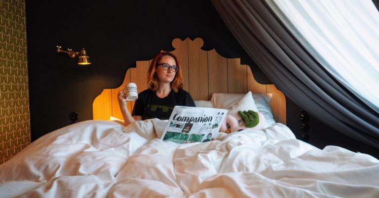 Sooz sitting in a bed with a white doona, reading magazine and holding a mug of coffee.