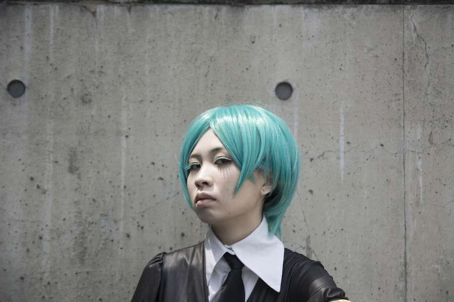 An Asian femme person wearing an aqua bob wig and a suit in front of a grey concrete wall.