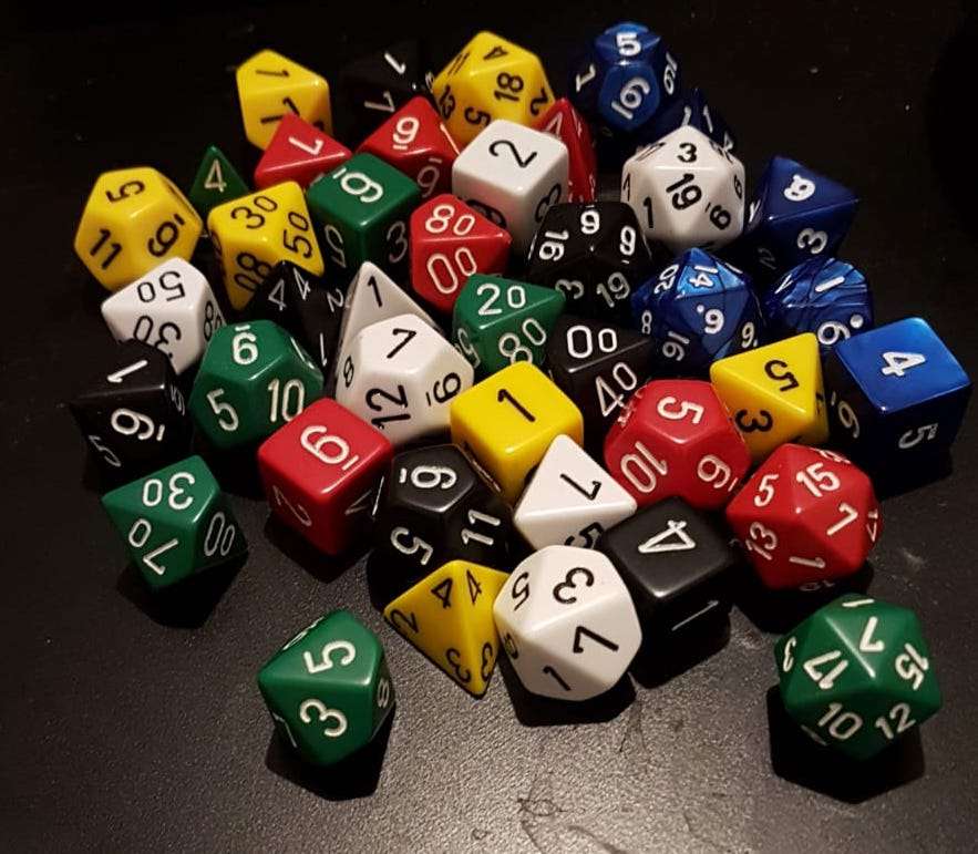 A close-up shot of a pile of different types of dice in red, white, black, green, blue and yellow on a black surface.