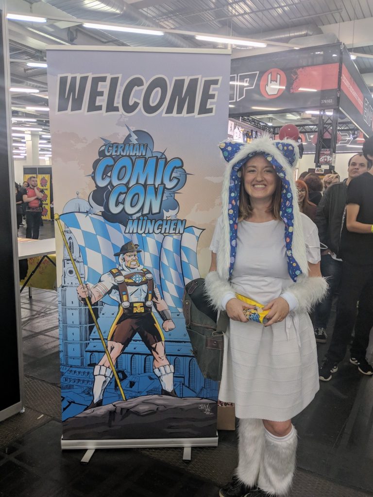 Sooz wearing a white costume and smiling at the camera while standing next to a sign saying "Welcome German Comic Con München."
