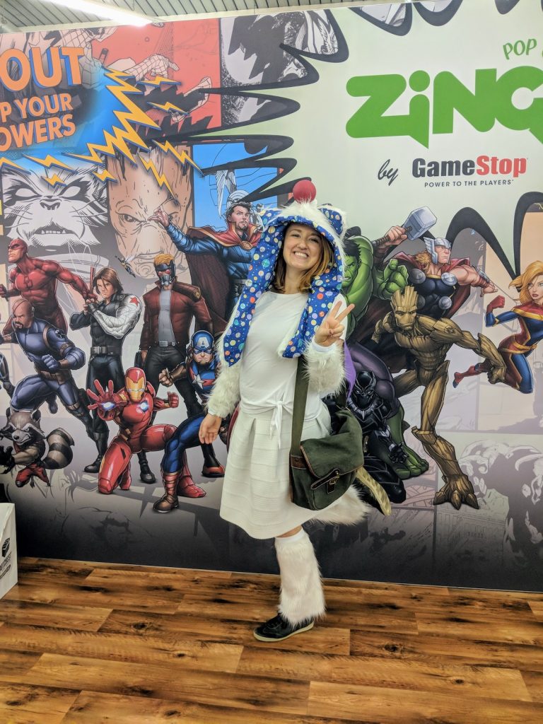 Sooz wearing a white costume and shoulder bag doing the peace sign with one leg in the air in front of a backdrop featuring different Marvel comic characters.