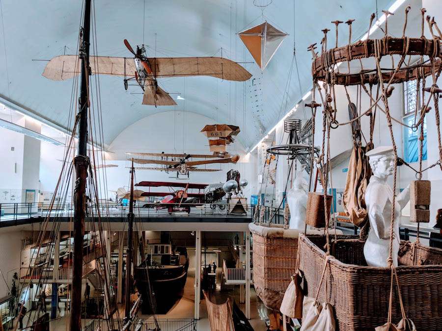 A high ceilinged room with old fashioned airplanes and hot air balloon baskets hanging down and ship masts pointing up from the lower floor.