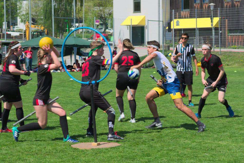 A group of people on a grass field holding sticks between their legs and playing a ball game in front of a ring on a pedestal.