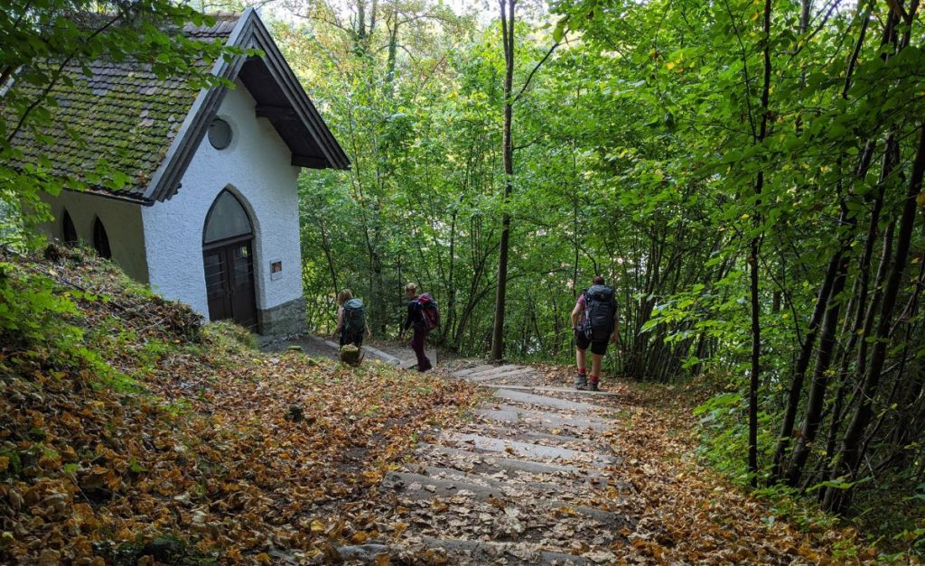 Three people wearing backpacks walking down stairs covered in autumn leaves to a small white building in a forest.
