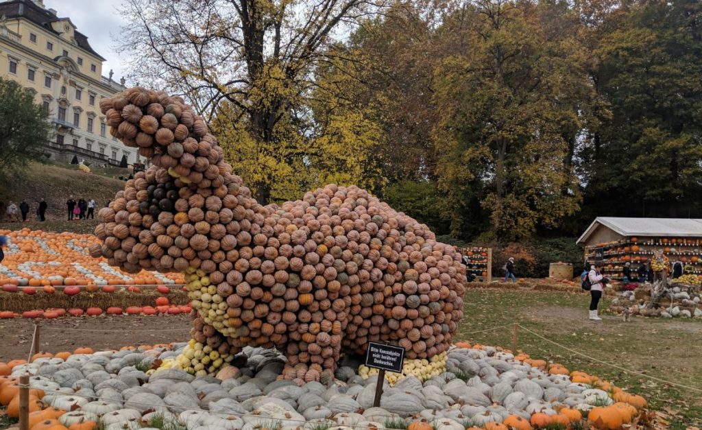 A giant rabbit sculpture made of pumpkins in the grounds before a cream colored mansion with trees, stalls and more pumpkins behind the sculpture.