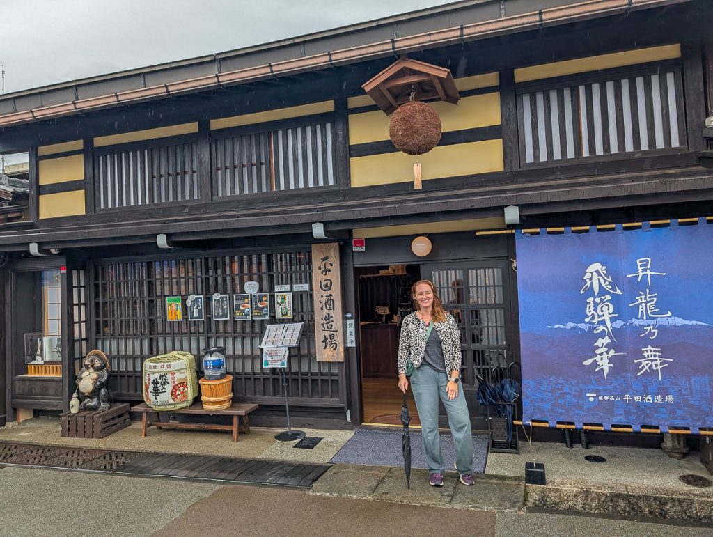 Sool holding a closed umbrella and standing in front of a sake brewery while smiling at the camera.
