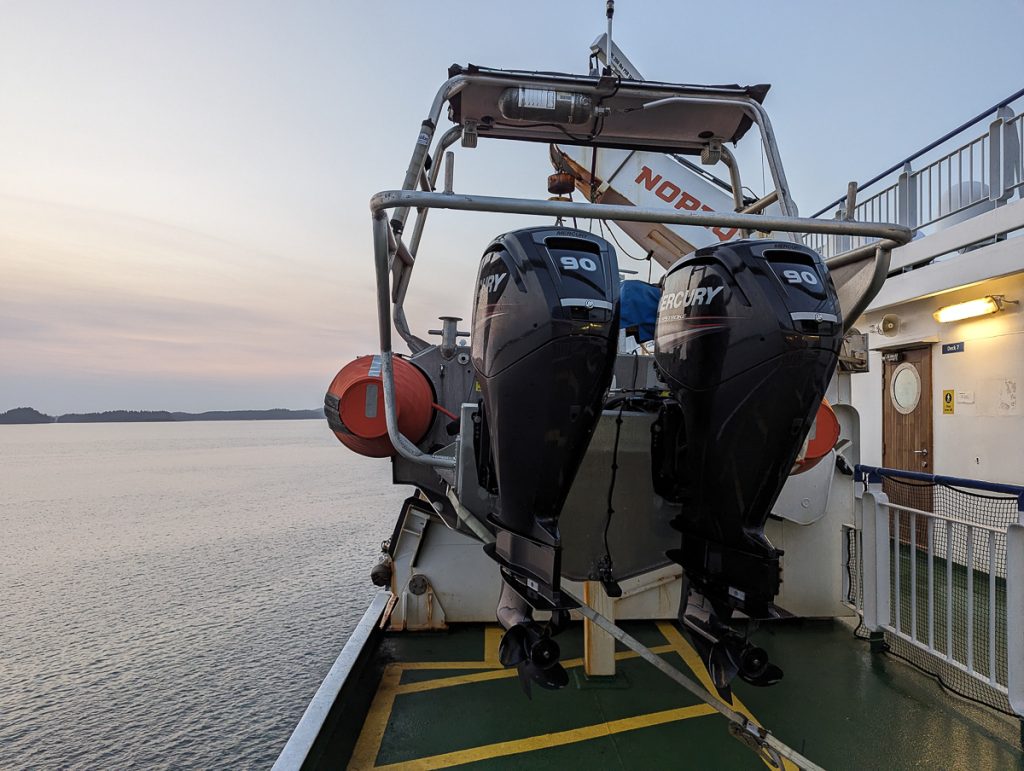 A smaller boat with outboard motors on the side of a larger ferry at dusk.