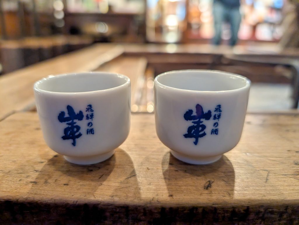 A close-up shot of two white sake cups, with blue Japanese writing on the sides.