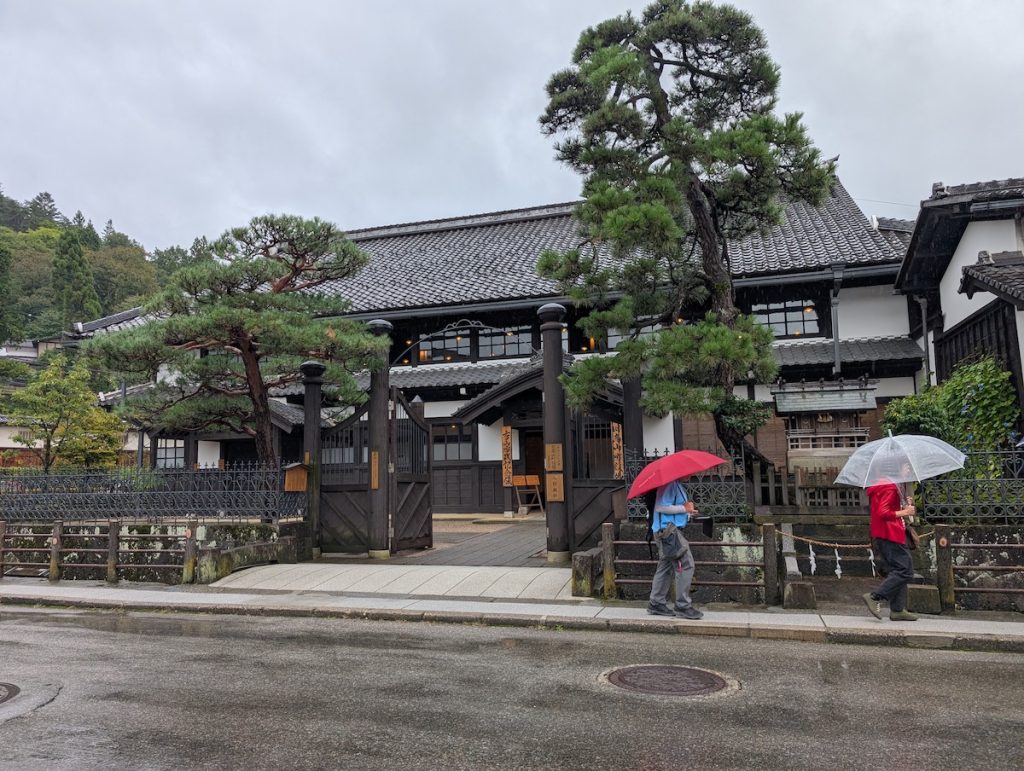 Two people holding umbrellas walking past a traditional building in Takayama on a rainy day.