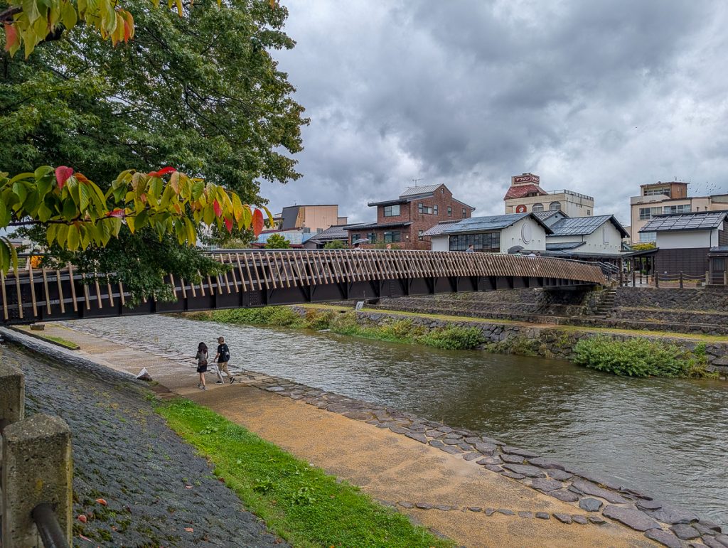 Two people walking under a bridge going over a river on a cloudy day.