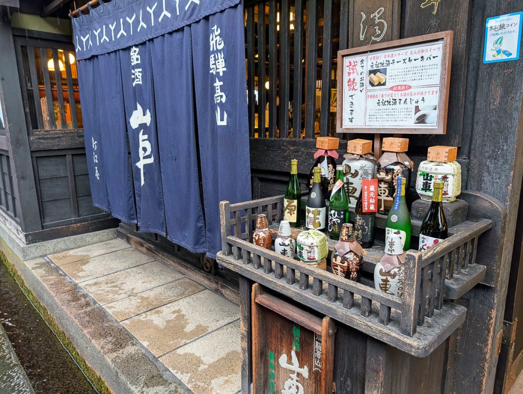 A wooden stand with bottles of different sake on display in front of a brewery.