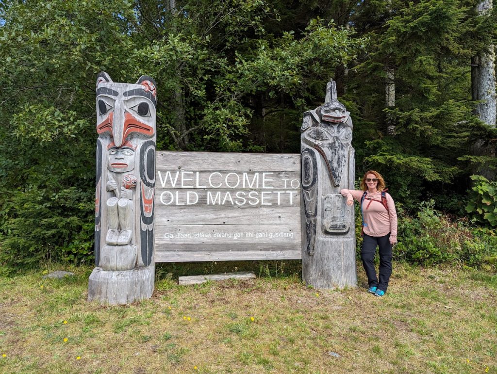 Sooz standing next to two small wooden totems and a sign that says "Welcome to Old Massett"" with Haida  language underneath.