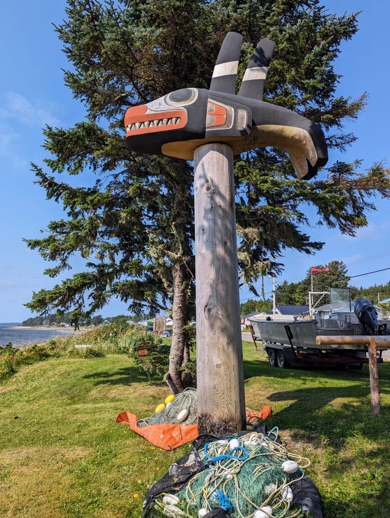A Haida carving of an orca on a wooden pole with fishnets on the grass beneath, a boat on a trailer behind and a tree and a beach in the background.