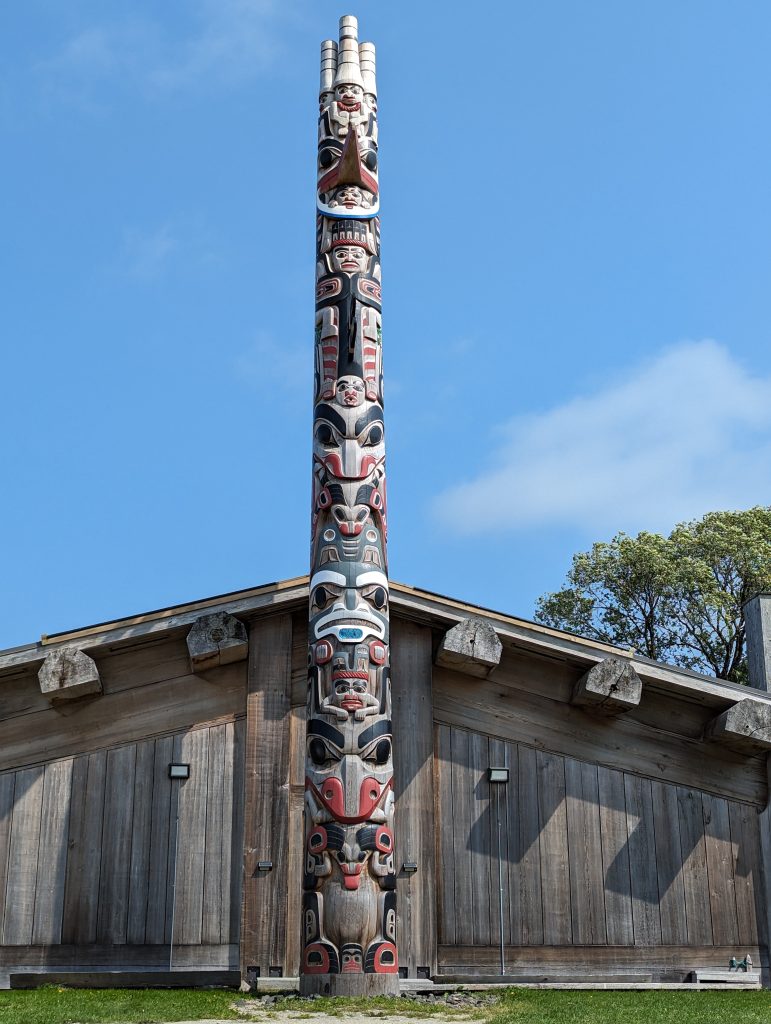 A Haida chief's totem pole in front of a wooden building and blue sky.