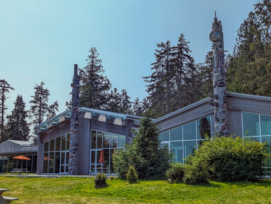 A building with big windows and two totem poles out the front, with trees and blue sky behind.