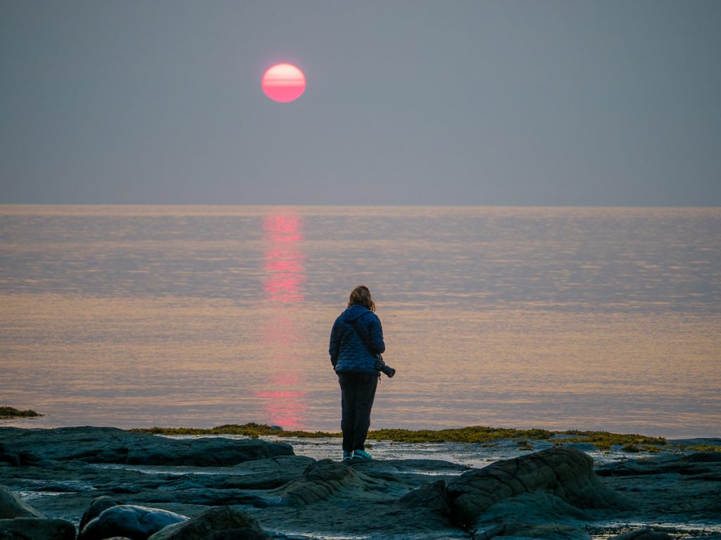 Sooz standing on rocks looking towards the ocean with an incredible pink moon rising in the sky.