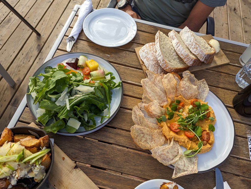 An overhead shot of a wooden table with plates of food including salad, bread and more yummy looking dishes.