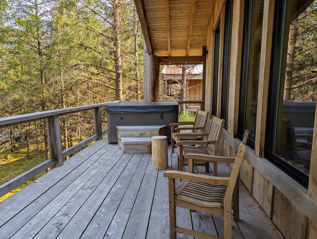 A wooden deck with wooden chairs and a hot tub overlooking pine trees.