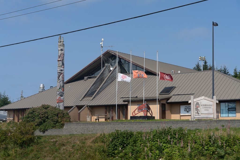 A building with a pointed roof with flags and a totem pole out the front with a sign saying "Kwiiyaans Community Hall."