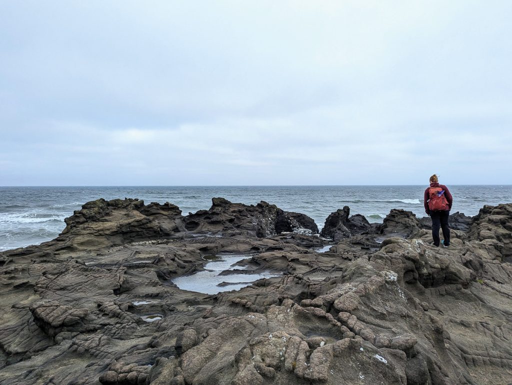 Sooz standing on rugged ocean rocks next to the water with an overcast sky.