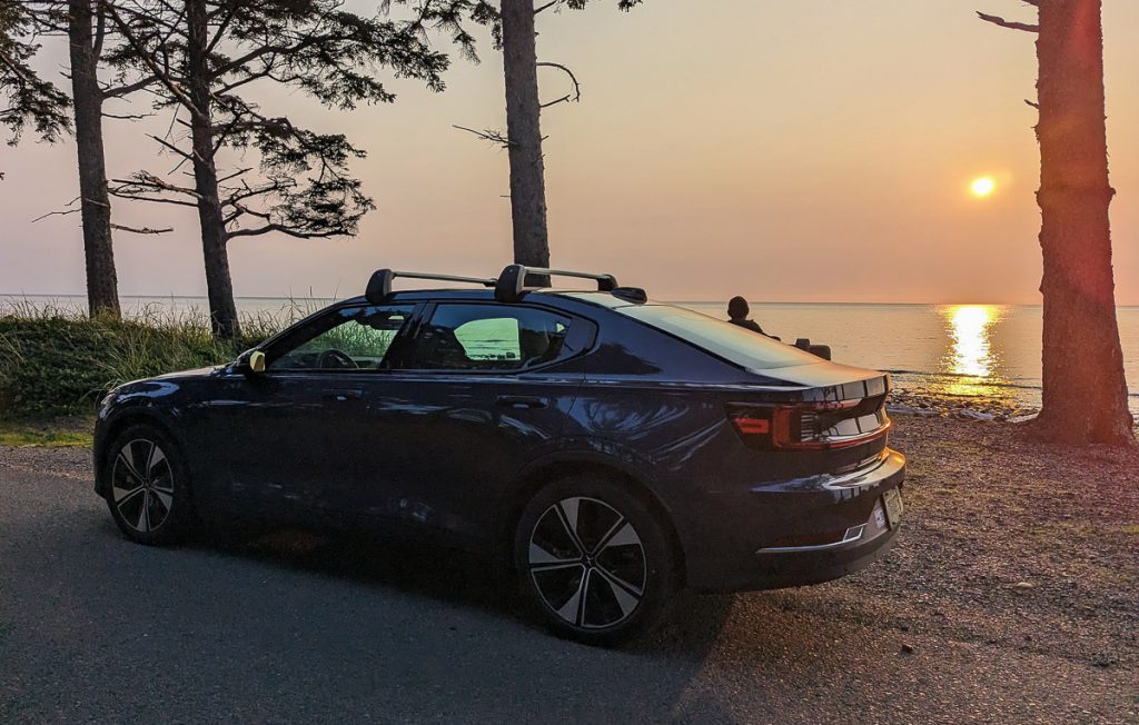 An electric car parked on the side of a ride directly next to a beach at sunset.